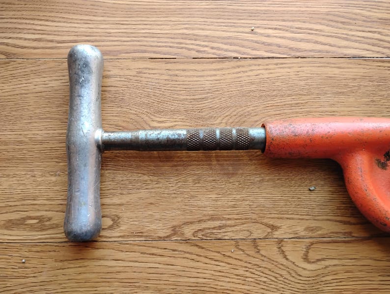 Pode incluir: Uma ferramenta de metal antiga com uma al&ccedil;a em forma de T e uma pega laranja. A al&ccedil;a &eacute; prateada e mostra sinais de desgaste. A pega laranja tem um design curvo. A ferramenta est&aacute; sobre um fundo de madeira.