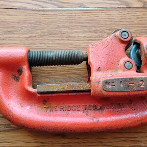 Pode incluir: Cortador de tubos laranja da The Ridge Tool Company. A ferramenta tem um corpo curvo com uma roda de corte e um mecanismo de ajuste roscado. O n&uacute;mero do modelo "No. 1-2" &eacute; vis&iacute;vel na ferramenta.