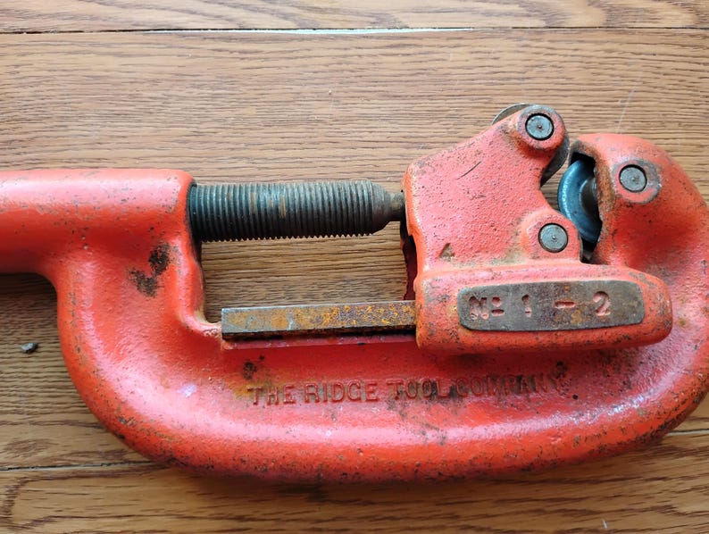 Pode incluir: Cortador de tubos laranja da The Ridge Tool Company. A ferramenta tem um corpo curvo com uma roda de corte e um mecanismo de ajuste roscado. O n&uacute;mero do modelo "No. 1-2" &eacute; vis&iacute;vel na ferramenta.