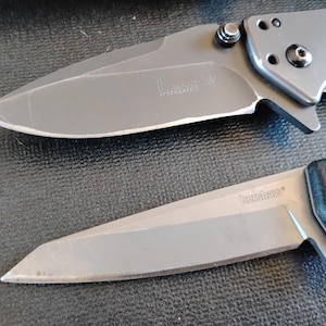 Puede incluir: Dos navajas plegables con hojas y mangos de metal gris. La navaja superior presenta una hoja curva con el texto Kershaw SpeedSafe. La navaja inferior tiene una hoja de borde recto con el texto Kershaw. Ambas hojas son de color gris met&aacute;lico.
