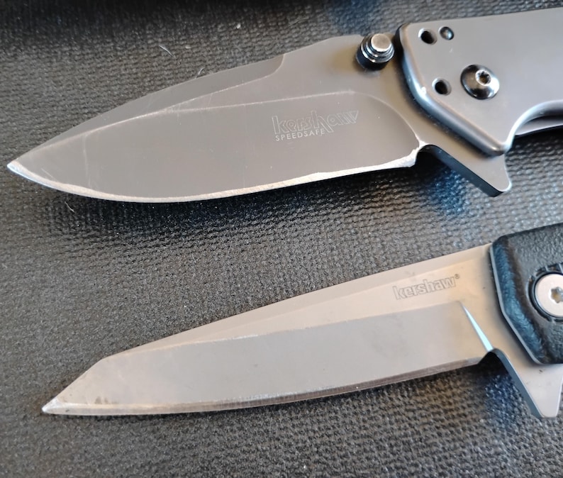 Puede incluir: Dos navajas plegables con hojas y mangos de metal gris. La navaja superior presenta una hoja curva con el texto Kershaw SpeedSafe. La navaja inferior tiene una hoja de borde recto con el texto Kershaw. Ambas hojas son de color gris met&aacute;lico.