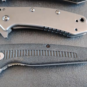 Puede incluir: Se muestran dos navajas plegables. La navaja superior presenta un mango de metal gris mate liso con tornillos visibles y un orificio para cord&oacute;n. La navaja inferior tiene un mango negro texturizado con un patr&oacute;n de ranuras central y un tornillo de pivote plateado.