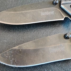 Puede incluir: Dos navajas plegables con hojas negras y mangos met&aacute;licos. Cada hoja presenta el nombre de la marca Kershaw y el texto SpeedSafe grabados en la superficie. Las navajas est&aacute;n colocadas horizontalmente, mostrando los filos y los pivotes.