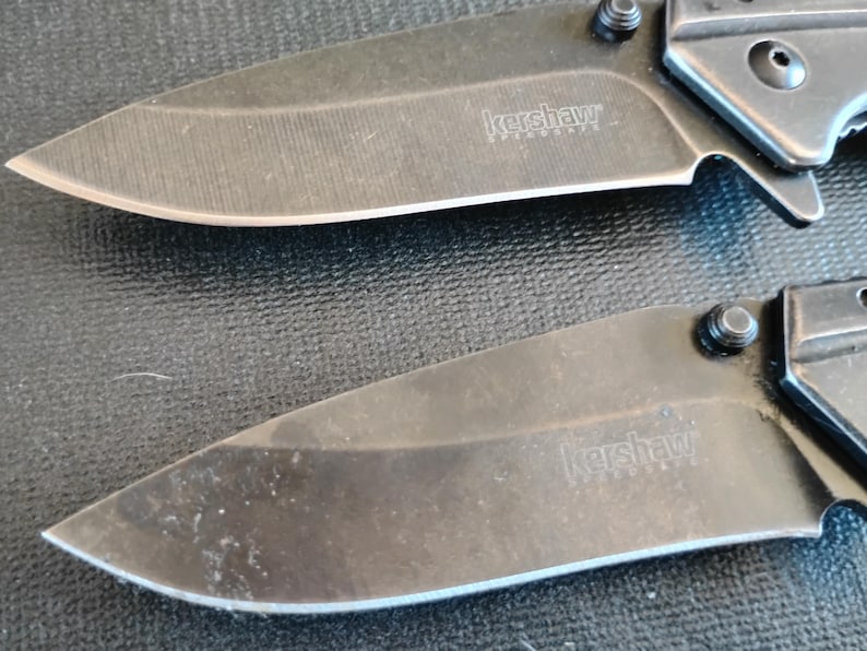 Puede incluir: Dos navajas plegables con hojas negras y mangos met&aacute;licos. Cada hoja presenta el nombre de la marca Kershaw y el texto SpeedSafe grabados en la superficie. Las navajas est&aacute;n colocadas horizontalmente, mostrando los filos y los pivotes.