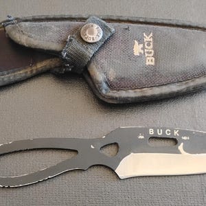 May include: A black Buck knife with a silver blade and a matching black sheath. The knife has a unique handle design with a hole. The sheath is made of a durable, textured material and features the Buck logo.