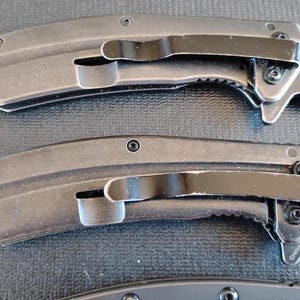 Puede incluir: Dos navajas plegables con mangos de metal gris oscuro y clips de bolsillo negros. Cada mango presenta un dise&ntilde;o curvo con tornillos visibles y un acabado texturizado. Las navajas est&aacute;n colocadas horizontalmente, mostrando el perfil lateral.