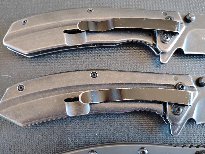 Puede incluir: Dos navajas plegables con mangos de metal gris oscuro y clips de bolsillo negros. Cada mango presenta un dise&ntilde;o curvo con tornillos visibles y un acabado texturizado. Las navajas est&aacute;n colocadas horizontalmente, mostrando el perfil lateral.