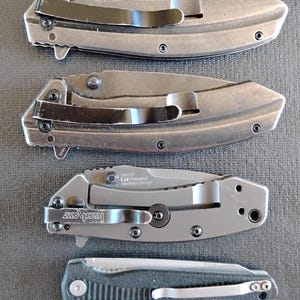 Puede incluir: Cuatro navajas plegables dispuestas verticalmente. Las dos superiores son grises con clips negros. La tercera es una Kershaw 1555TI Hinderer Design gris con clip negro. La navaja inferior es negra con mango texturizado y clip plateado.
