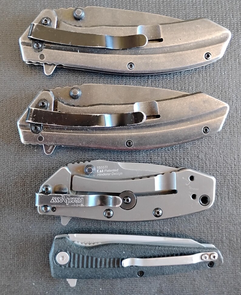 Puede incluir: Cuatro navajas plegables dispuestas verticalmente. Las dos superiores son grises con clips negros. La tercera es una Kershaw 1555TI Hinderer Design gris con clip negro. La navaja inferior es negra con mango texturizado y clip plateado.