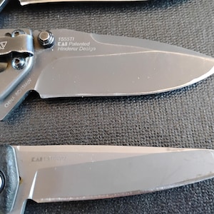 Puede incluir: Dos navajas plegables con hojas y mangos de metal gris. La navaja superior tiene un clip de bolsillo y texto en la hoja que dice 1555TI KAI Patented Hinderer Design y CHINA 8Cr13MoV. La navaja inferior tiene un mango negro con textura.
