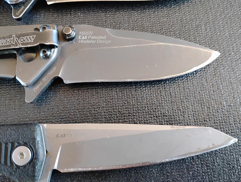 Puede incluir: Dos navajas plegables con hojas y mangos de metal gris. La navaja superior tiene un clip de bolsillo y texto en la hoja que dice 1555TI KAI Patented Hinderer Design y CHINA 8Cr13MoV. La navaja inferior tiene un mango negro con textura.