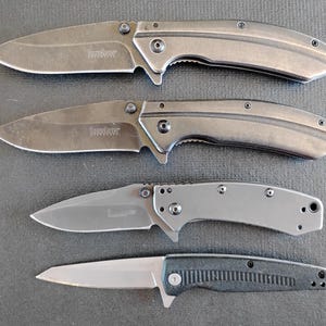 Puede incluir: Cuatro navajas plegables dispuestas verticalmente. Las dos superiores son de color gris oscuro con mangos curvos y el logotipo de Kershaw. La tercera es de un gris m&aacute;s claro con mango liso. La navaja inferior tiene una hoja plateada y un mango negro texturizado.