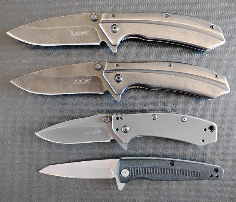 Puede incluir: Cuatro navajas plegables dispuestas verticalmente. Las dos superiores son de color gris oscuro con mangos curvos y el logotipo de Kershaw. La tercera es de un gris m&aacute;s claro con mango liso. La navaja inferior tiene una hoja plateada y un mango negro texturizado.
