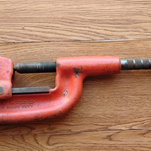 Pode incluir: Um cortador de tubos laranja e prateado, com a marca "RIDGID" vis&iacute;vel. A ferramenta tem uma pega em forma de T e um mecanismo de ajuste roscado. A ferramenta foi concebida para utiliza&ccedil;&atilde;o intensiva.