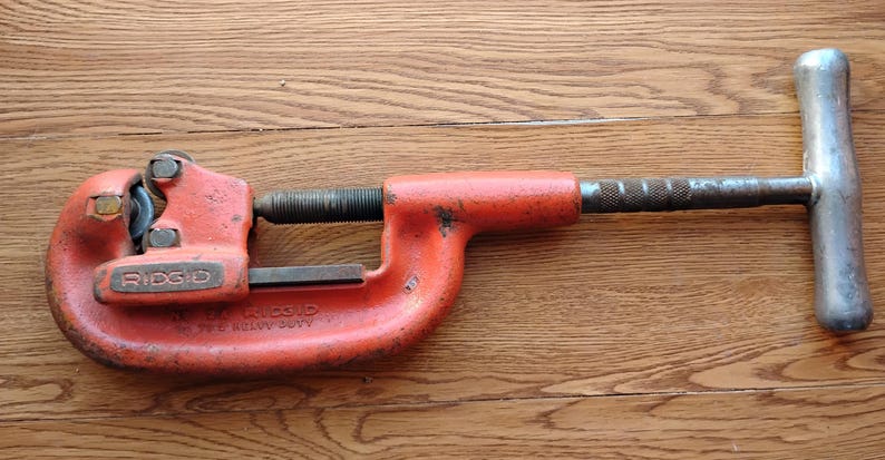 Pode incluir: Um cortador de tubos laranja e prateado, com a marca "RIDGID" vis&iacute;vel. A ferramenta tem uma pega em forma de T e um mecanismo de ajuste roscado. A ferramenta foi concebida para utiliza&ccedil;&atilde;o intensiva.