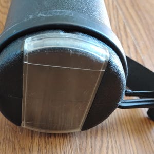 May include: Close-up of a black cylindrical object with a clear plastic cover. A black strap and buckle are attached to the object. The object appears to be made of a textured material.