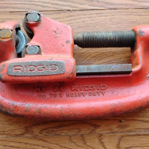 Pode incluir: Um cortador de tubos Ridgid vintage e resistente, pintado em laranja desgastado. A ferramenta possui um mecanismo de parafuso e uma roda de corte. O texto "RIDGID" e "NO. 24" s&atilde;o vis&iacute;veis na ferramenta.