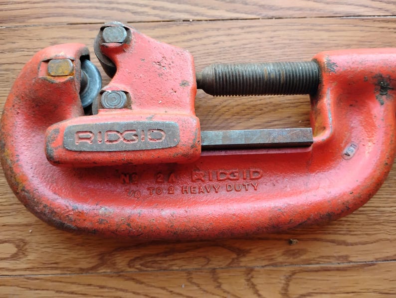 Pode incluir: Um cortador de tubos Ridgid vintage e resistente, pintado em laranja desgastado. A ferramenta possui um mecanismo de parafuso e uma roda de corte. O texto "RIDGID" e "NO. 24" s&atilde;o vis&iacute;veis na ferramenta.