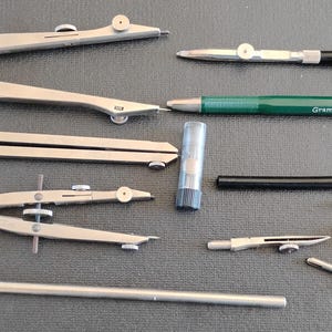 May include: An assortment of drafting tools, including several compasses, a green and silver mechanical pencil with the text "Gramercy - ITALY - 2100", and various other drawing instruments. The tools are silver and black, arranged on a dark gray surface.