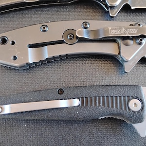 Puede incluir: Dos navajas plegables con mangos de metal. La navaja superior es gris con un clip de bolsillo negro que muestra el logotipo de Kershaw. La navaja inferior tiene un mango negro texturizado y un clip de bolsillo plateado. Ambas tienen tornillos.