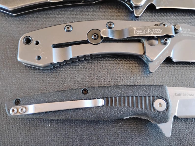 Puede incluir: Dos navajas plegables con mangos de metal. La navaja superior es gris con un clip de bolsillo negro que muestra el logotipo de Kershaw. La navaja inferior tiene un mango negro texturizado y un clip de bolsillo plateado. Ambas tienen tornillos.