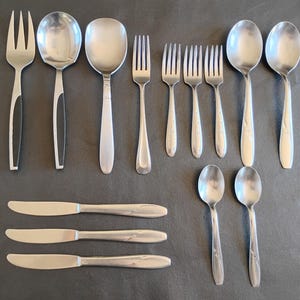 May include: A collection of twelve stainless steel flatware pieces including three dinner knives, three dinner forks, three dinner spoons, two smaller spoons, and one serving fork and spoon set with black accents on the handles of the larger pieces.