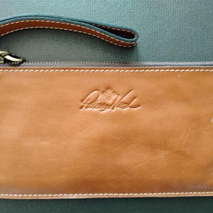 May include: A brown leather clutch with a wrist strap and a zipper closure. The clutch features white stitching and an embossed logo. The wrist strap is brown with a dark blue interior. The clutch is approximately 9 inches long.