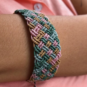 Puede incluir: Una pulsera de macramé hecha a mano con un patrón de chevrón. La pulsera presenta una combinación de hilos verdes, rosas y beige. La pulsera se lleva en la muñeca, y el fondo es una camisa rosa claro.