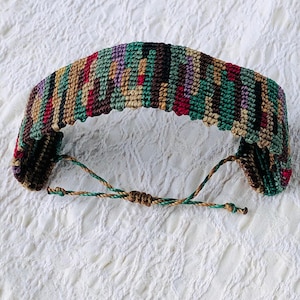 May include: A handmade macrame bracelet featuring a woven pattern in shades of green, brown, red, and purple. The bracelet has adjustable drawstrings for a custom fit. The intricate design and earthy tones make it a unique accessory.