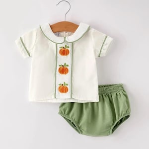 May include: A two-piece outfit consisting of a short-sleeved white shirt with a Peter Pan collar, green piping, and a vertical center panel featuring three embroidered orange pumpkins, paired with solid sage green elastic-waist bloomers.