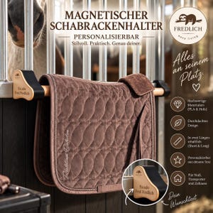 Op de afbeelding: Een bruin gewatteerd zadeldekje hangt aan een houten stang met zwarte magnetische houders. De houders zijn gepersonaliseerd met de tekst Escado Fred Fredlich. De afbeelding bevat tekst zoals MAGNETISCHER SCHABRACKENHALTER en FREDLICH.