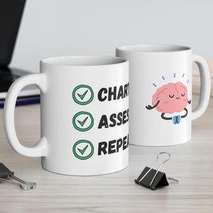 May include: Two white ceramic mugs. One mug has the words "CHAR, ASSES, REPE" with checkmarks. The other mug features a cartoon brain with arms and legs. The brain is pink with a smiling face and is meditating.