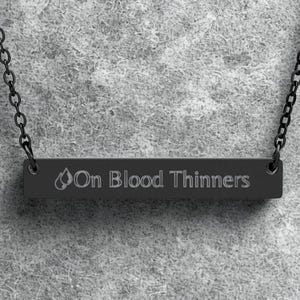 Puede incluir: Collar negro con un colgante rectangular con la inscripción grabada "On Blood Thinners" y un símbolo de llama. La cadena y el colgante son negros.