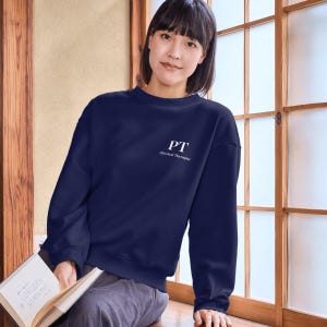 Puede incluir: Sudadera azul marino con las letras blancas "PT" y las palabras "Physical Therapist". La sudadera es de cuello redondo y está hecha de un material suave. La persona de la imagen sostiene un libro.