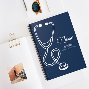 Puede incluir: Un diario de espiral azul marino con un diseño de estetoscopio blanco. La portada dice "Nurse Journal" con las palabras "Breathe, Care, Heal" debajo. El diario está sobre una superficie blanca.