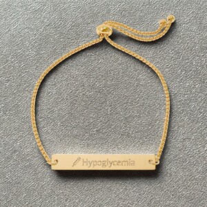 Op de afbeelding: Een goudkleurige verstelbare armband met een rechthoekige hanger. De hanger is gegraveerd met het woord "Hypoglycemie" en een klein spuitpictogram. De armband heeft een fijne ketting en een schuifkralensluiting.