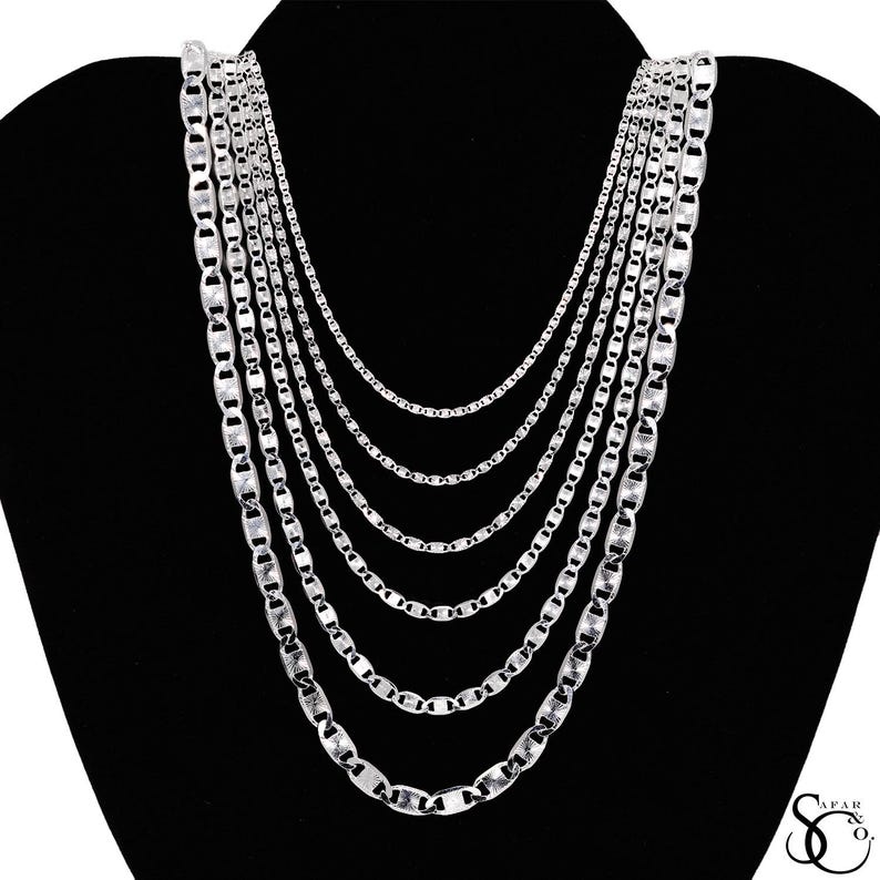 May include: A collection of silver necklaces displayed on a black mannequin. The necklaces vary in length and chain style, creating a layered effect. The chains have a shiny, reflective surface, and the background is a solid black.