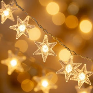 May include: A string of clear, star-shaped fairy lights with warm white LED bulbs. The lights are strung on a thin, silver wire, creating a festive and decorative ambiance. The background is a soft, blurred golden color.