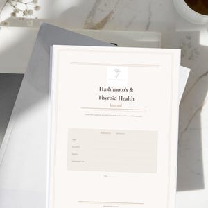 Puede incluir: Un diario blanco titulado "Hashimoto's & Thyroid Health Journal" con un logotipo floral. El diario está abierto en una página con secciones rayadas para notas. El diario está sobre una pila de libros, con una taza de café en el fondo.