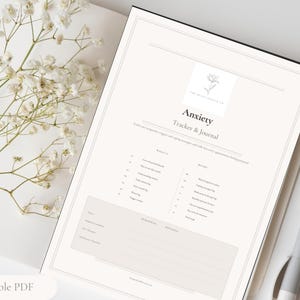May include: A printable PDF anxiety tracker and journal with a floral logo. The journal is open to a page with the title "Anxiety Tracker & Journal". A white pen rests on the right side of the journal. White flowers are in the upper left corner.