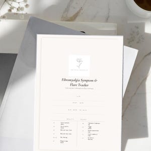 Op de afbeelding: Een crèmekleurige Fibromyalgie Symptoom & Flare Tracker, met een bloemenlogo, ligt bovenop een stapel witte en grijze papieren. Een kopje koffie en takjes witte bloemen staan op de achtergrond. De tracker bevat secties voor het volgen van symptomen en flares.