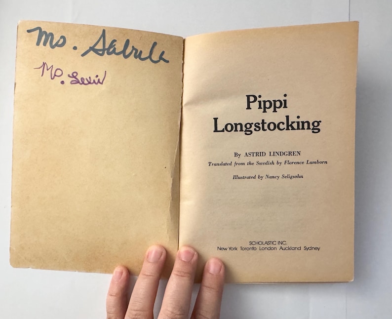 Puede incluir: Un libro abierto con el t&iacute;tulo "Pippi Longstocking" de Astrid Lindgren. Las p&aacute;ginas est&aacute;n envejecidas y tienen notas manuscritas. La portada muestra el logotipo de Scholastic Inc. y los lugares de publicaci&oacute;n.