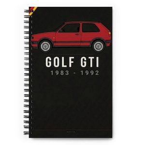 May include: A spiral-bound notebook with a black cover featuring a red car illustration. The text "GOLF GTI 1983 - 1992" is printed in white. A German flag design is on the top left corner.