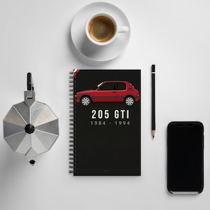May include: A black spiral-bound notebook featuring a red illustration of a 205 GTI car with the text "205 GTI 1984 - 1994". The image also includes a cup of coffee, a pencil, a smartphone, and a silver coffee maker.