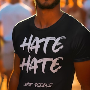 May include: Black crew neck t-shirt with the words "HATE HATE ...NOT PEOPLE!" in white brushstroke lettering. The shirt is worn by a person in a crowd, with blurred figures in the background.