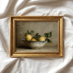 Op de afbeelding: Een ingelijst olieverfschilderij met een stilleven van drie gele citroenen met groene bladeren in een ondiepe, wit-blauwe keramische kom. Het kunstwerk is gevat in een decoratieve, textuurrijke goudkleurige lijst met een klassiek profiel.