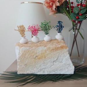 May include: A decorative arrangement featuring small white vases with colorful dried flowers atop a textured stone. A larger glass vase holds a bouquet of mixed flowers and berries. The scene is set on a wooden surface.