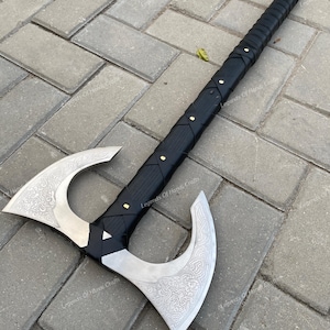 May include: A double-bit battle axe featuring a long black handle wrapped in leather with brass rivets. The two silver-coloured metal axe heads are decorated with intricate, etched knotwork patterns on both sides of the symmetrical blades.