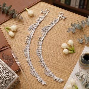 May include: A pair of silver-toned anklets featuring an intricate, scalloped lace-like design with small dangling charms along the edges. Each anklet is finished with a secure hook-and-eye clasp mechanism at the top for easy fastening.