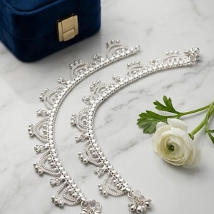 May include: A pair of silver-toned anklets featuring a delicate chain with repeating arched filigree patterns, small spherical beads, and dangling bell-shaped charms along the lower edge, arranged in a curved formation on a white surface.